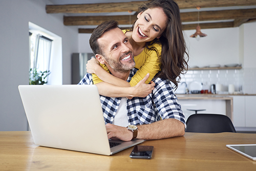 Joyful couple using a laptop at the dining table, smiling and collaborating